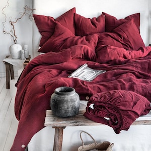 Puede incluir: Una funda nórdica y fundas de almohada de lino color burdeos sobre una cama blanca con cabecero de madera. La ropa de cama está dispuesta en un estilo relajado y casual. Un taburete de madera con una maceta de cerámica se encuentra delante de la cama.