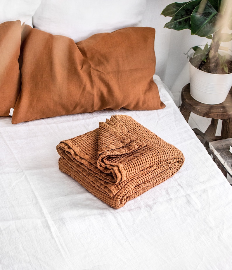 Pu&ograve; includere: Una coperta in tessuto a nido d'ape piegata in un caldo colore marrone su un letto di lino bianco. La coperta &egrave; realizzata in fibre naturali e ha una superficie morbida e testurizzata.