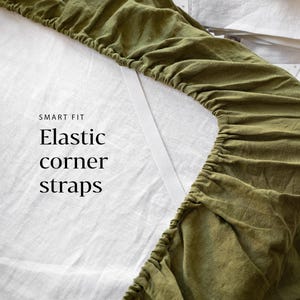 Op de afbeelding: Close-up van een olijfgroen hoeslaken met elastische hoekbanden. Het laken ligt op een wit bed. De tekst "SMART FIT" en "Elastic corner straps" zijn op het laken gedrukt.