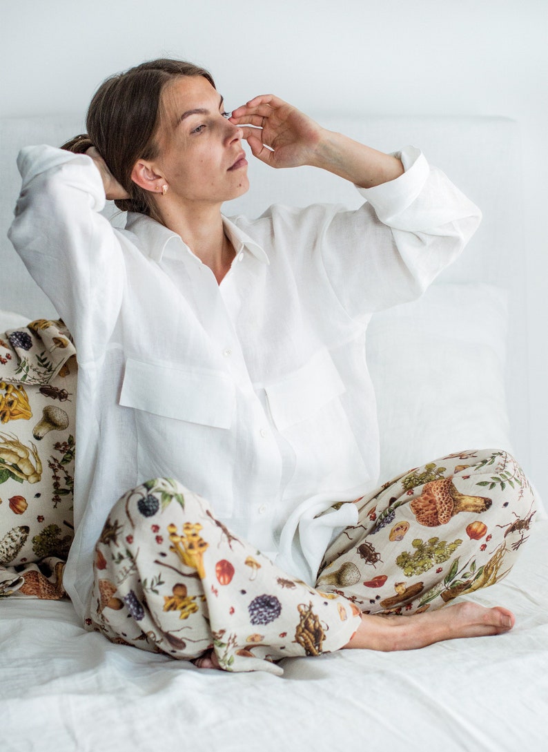 May include: A woman wearing a white linen shirt and trousers with a mushroom and insect print. She is sitting on a white bed with a pillow featuring a similar print.