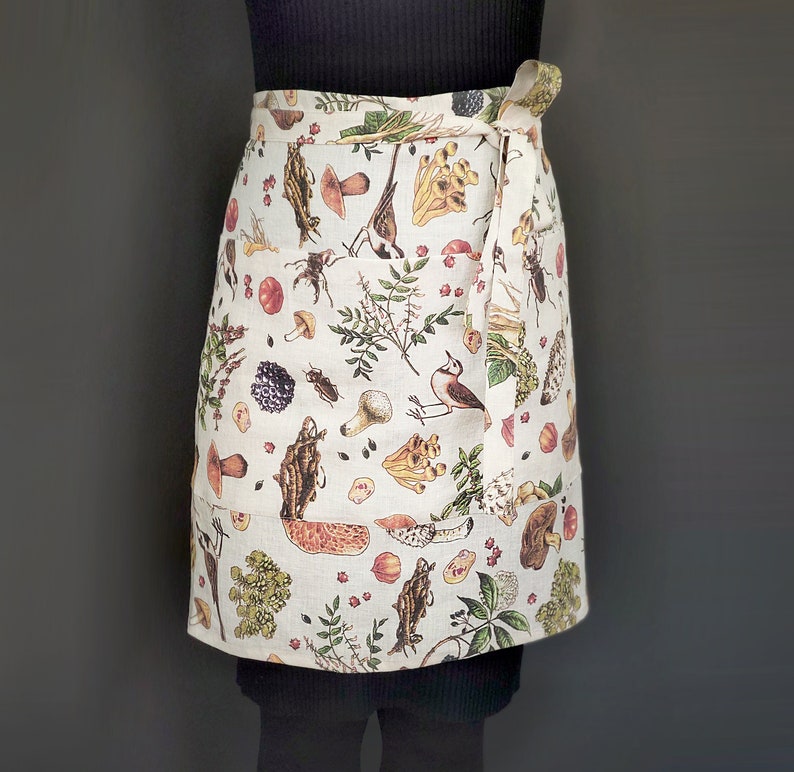 May include: A cream-colored apron with a wrap-around design, featuring a botanical print of mushrooms, berries, insects, and birds. The apron has a pocket on the front and a tie closure. The fabric appears to be linen or a similar natural material.