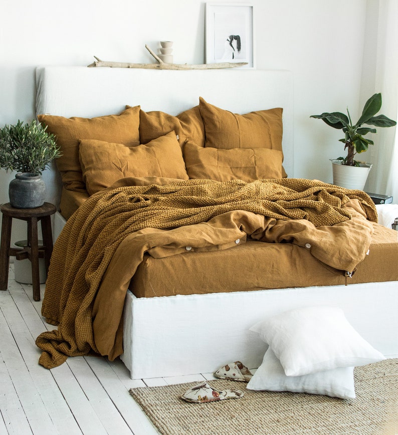 Puede incluir: Un juego de cama de lino amarillo mostaza con una manta de tejido de gofre. La ropa de cama est&aacute; sobre un marco de cama blanco con una cabecera blanca. Hay dos almohadas blancas en el suelo frente a la cama.