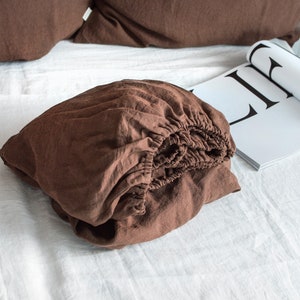 May include: A brown linen fitted sheet is folded and placed on a white linen bedspread.