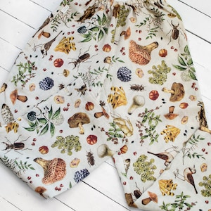 May include: A pair of beige linen trousers with a mushroom, bird, and insect print. The trousers have an elastic waistband and a relaxed fit.