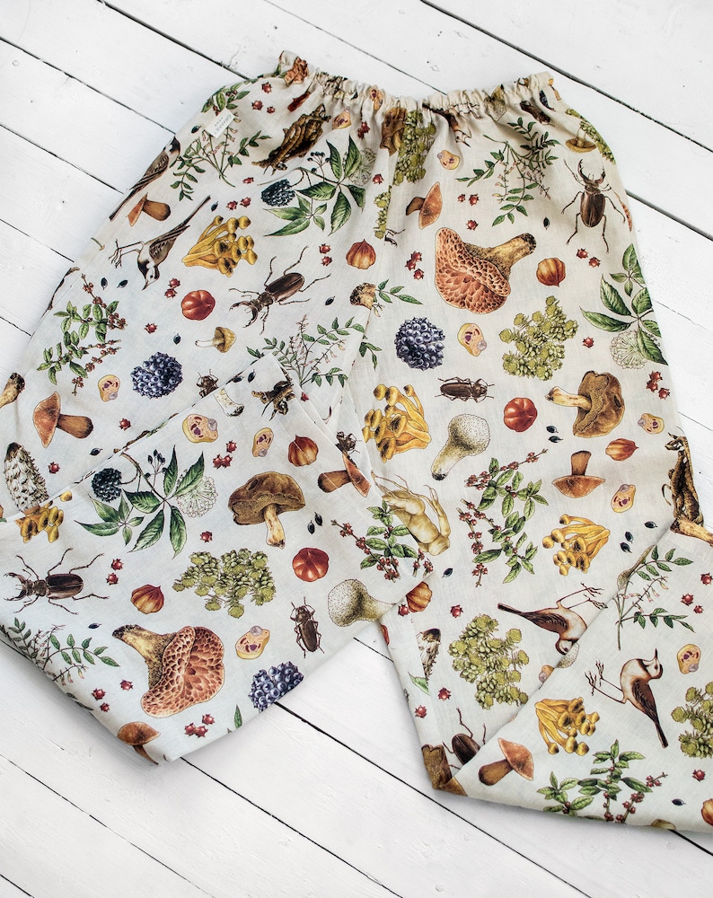 May include: A pair of beige linen trousers with a mushroom, bird, and insect print. The trousers have an elastic waistband and a relaxed fit.