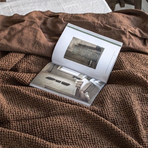 May include: A brown waffle weave throw blanket is folded on a bed with a white linen sheet and a brown linen pillow. A magazine is open on top of the blanket.