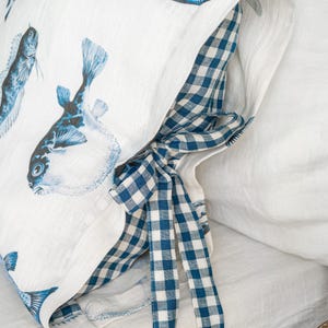 May include: White pillowcase with blue fish illustrations. The pillowcase has a blue and white gingham check pattern on the inside and a matching bow. The pillowcase is made of a soft, textured fabric.
