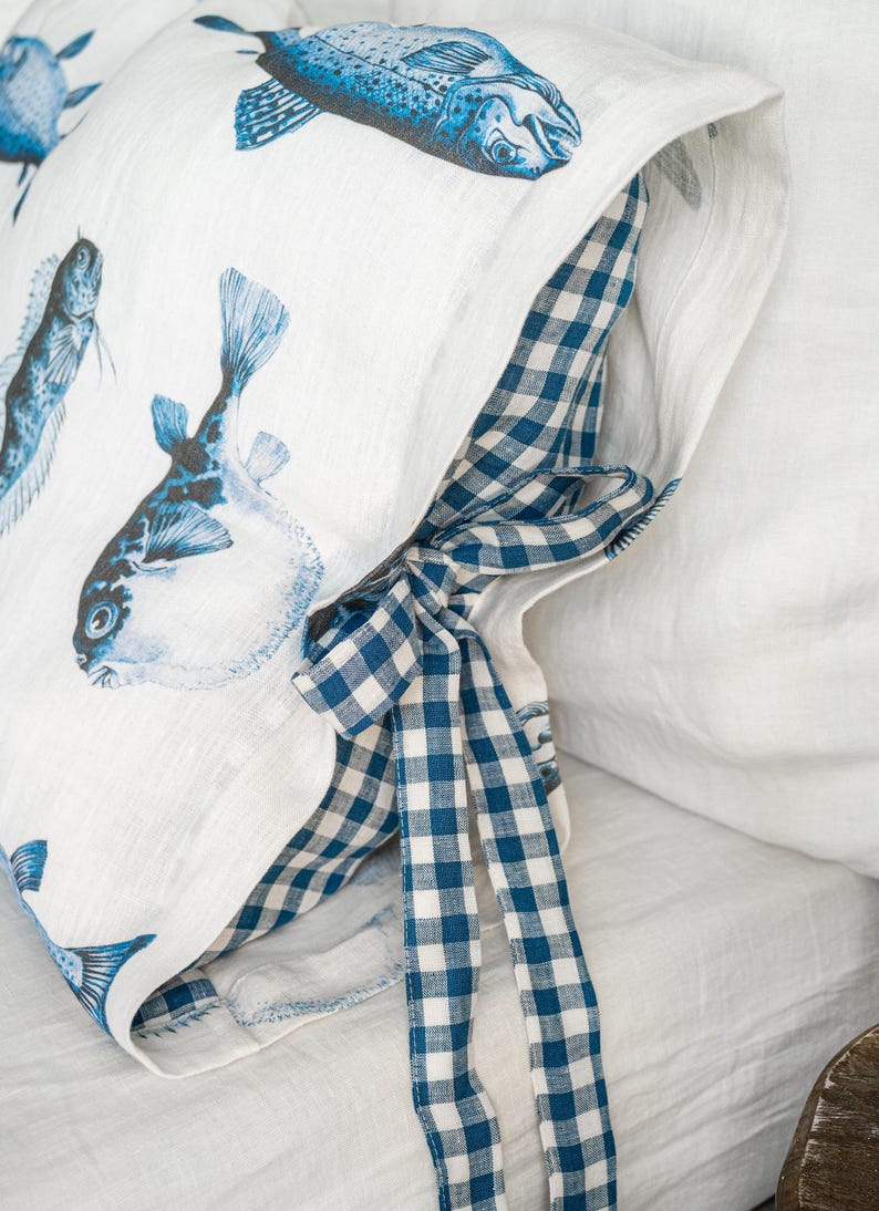 May include: White pillowcase with blue fish illustrations. The pillowcase has a blue and white gingham check pattern on the inside and a matching bow. The pillowcase is made of a soft, textured fabric.