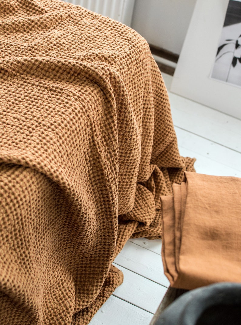 Pu&ograve; includere: Una coperta di tessuto a nido d'ape marrone piegata su una superficie di legno bianco. La coperta &egrave; realizzata in un tessuto morbido e testurizzato.