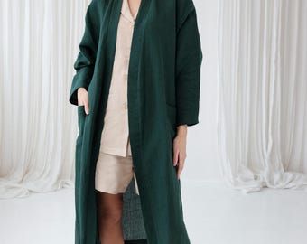 May include: A long, dark green linen robe with a shawl collar and pockets. The robe is open in the front, revealing a light beige top underneath.
