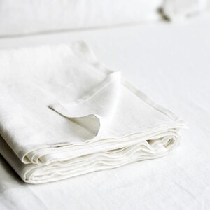 Peut inclure: Un empilement plié de linge de lit en lin blanc, possiblement un drap ou une couverture, est au centre de l'image. Le tissu semble doux et légèrement texturé, avec un éclat subtil. Le linge de lit est plié avec soin, une partie de la couche supérieure repliée.