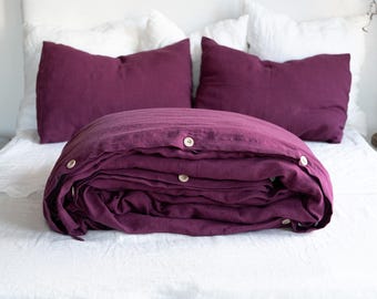 May include: A folded burgundy linen duvet cover with wooden buttons on a white bed. Three small concrete candles with the word "Harmony" on them are on a gray tray in the foreground.