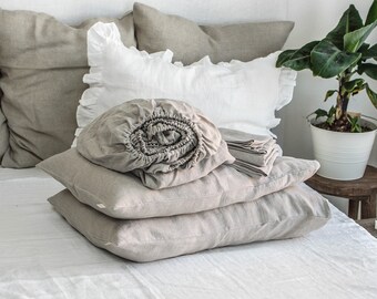 Puede incluir: Una pila de fundas de almohada y sábanas de lino gris claro sobre una cama blanca. El lino está doblado cuidadosamente y la tela tiene un aspecto suave y texturizado.