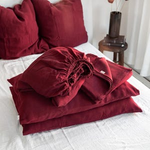 Puede incluir: Una pila de ropa de cama de lino roja, que incluye una sábana bajera, una sábana plana y fundas de almohada. La ropa de cama está dispuesta sobre un cubrecama blanco.