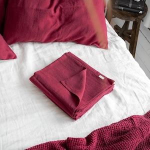 May include: A white linen bed with a folded burgundy linen throw blanket on top. A burgundy waffle weave throw blanket is at the bottom of the bed.