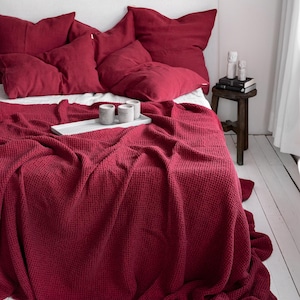 Puede incluir: Una manta de tejido de waffle rojo drapeada sobre una cama con almohadas de lino rojo. La manta está hecha de una tela suave y texturizada y tiene un aspecto relajado y casual.