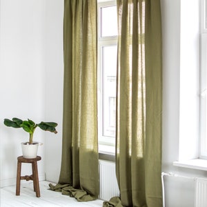 May include: Two olive green linen curtains hang in a window, with the curtains pulled back to reveal a white window frame and a view of a building outside. The curtains are gathered at the top and hang straight down to the floor.