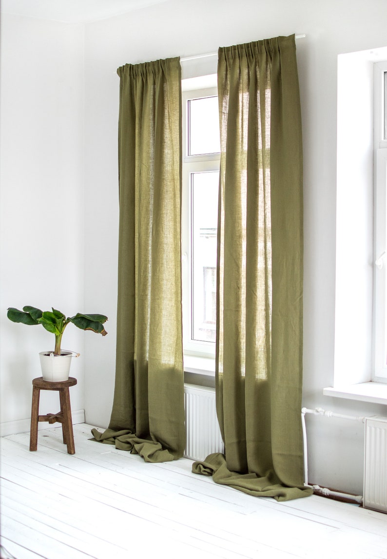 May include: Two olive green linen curtains hang in a window, with the curtains pulled back to reveal a white window frame and a view of a building outside. The curtains are gathered at the top and hang straight down to the floor.