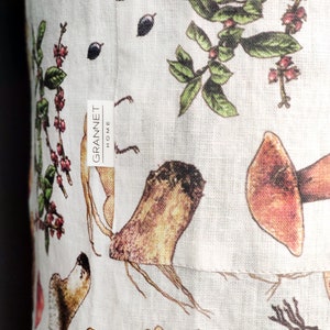 May include: Close-up of a white linen fabric with a botanical print featuring mushrooms, berries, and leaves in shades of brown, red, and green. The fabric has a tag that reads "GRANNET HOME".