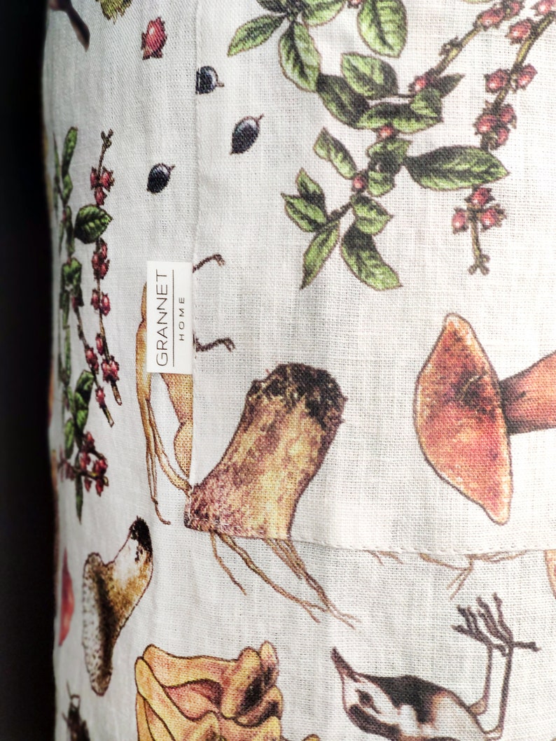 May include: Close-up of a white linen fabric with a botanical print featuring mushrooms, berries, and leaves in shades of brown, red, and green. The fabric has a tag that reads "GRANNET HOME".