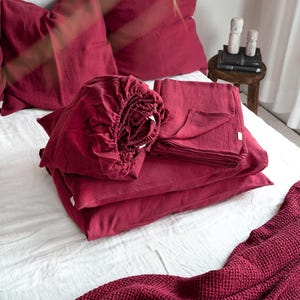 May include: A set of burgundy linen bedding, including a fitted sheet, flat sheet, pillowcases, and a duvet cover. The bedding is folded neatly on a white bed.