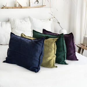 Pode incluir: Uma coleção de cinco almofadas de linho em várias cores: azul marinho, verde azeitona, verde escuro e roxo. As almofadas estão dispostas numa cama branca, criando um ambiente acolhedor. Almofadas decorativas para o quarto.