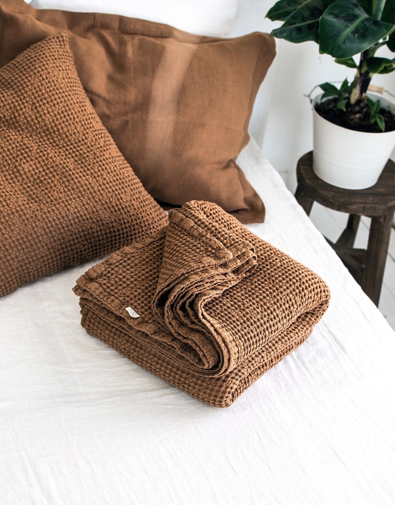 May include: A brown waffle weave throw blanket folded on a white bed with a brown waffle weave pillow on top of the bed.