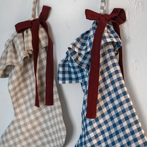 May include: Two Christmas stockings with ruffled tops and gingham patterns. One stocking is beige and white, the other is blue and white. Both have a burgundy ribbon bow and hanging loop.