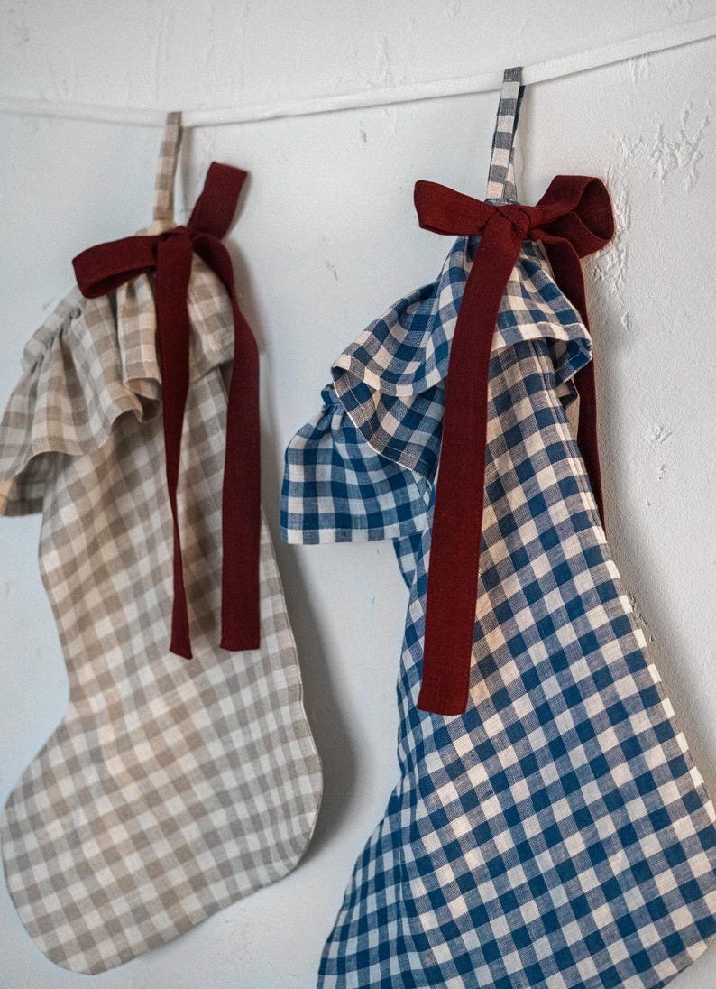 May include: Two Christmas stockings with ruffled tops and gingham patterns. One stocking is beige and white, the other is blue and white. Both have a burgundy ribbon bow and hanging loop.