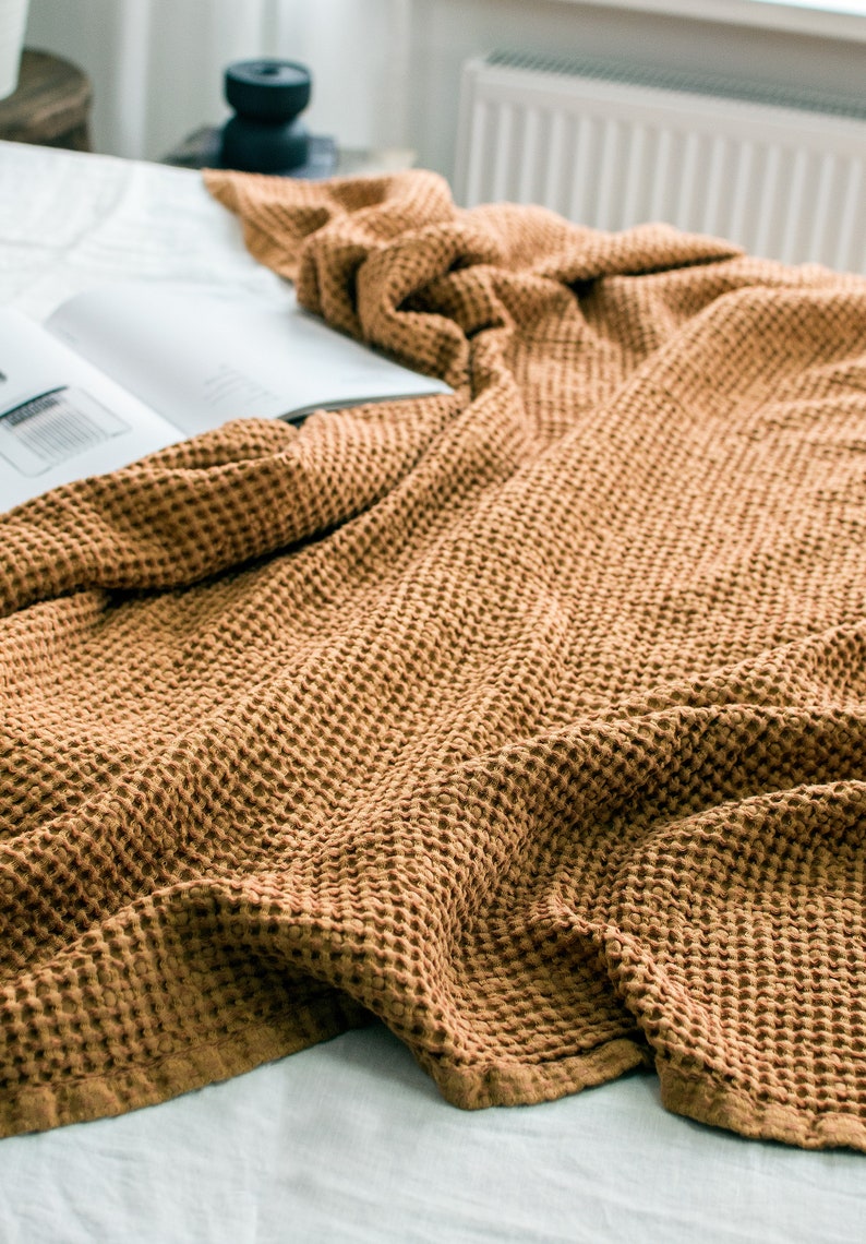 Pu&ograve; includere: Una coperta in tessuto waffle marrone &egrave; drappeggiata su un letto bianco. La coperta &egrave; realizzata in un tessuto morbido e testurizzato con un leggero bagliore.