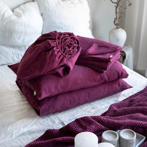 May include: A stack of burgundy linen bedding, including a fitted sheet, flat sheet, and pillowcases. The bedding is arranged on a white bed with a burgundy waffle weave throw blanket and white candles on a tray.