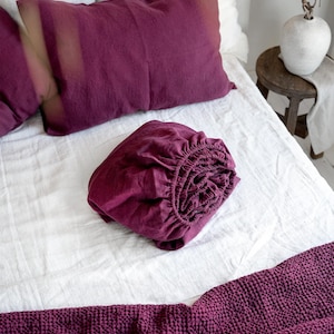 Op de afbeelding: Een opgerold bordeauxkleurig linnen hoeslaken op een wit bed met een bordeauxkleurig wafeldekentje aan het voeteneinde van het bed.