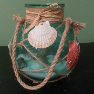 May include: A decorative glass jar with a teal interior, adorned with rope, seashells, and a raffia bow. The jar is wrapped in a rope net and features a large white seashell and a red seashell. The jar is filled with sand and small stones.