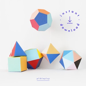 Könnte beinhalten: Ein Set aus sechs bunten geometrischen Papierformen, darunter eine Kugel, eine Pyramide und ein Würfel. Die Formen hängen mit einer Schnur von der Decke. Der Text "Sofort-Download" ist in einem Kreis mit einem nach unten zeigenden Pfeil und einer Wellenlinie geschrieben.