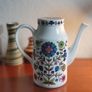 May include: White ceramic coffee pot with a floral design in shades of blue, green, pink, and brown. The pot has a spout and a handle.