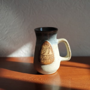 May include: A ceramic mug with a brown and white glaze. The mug has a handle and a brown design that says "1977 Silver Jubilee" with a crown above the year.