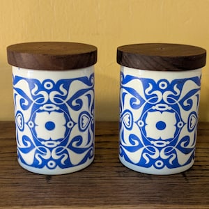 May include: Two matching salt and pepper shakers. Each has a white ceramic body with a blue floral design and a dark brown wooden lid. The shakers are cylindrical and sit on a wooden surface.