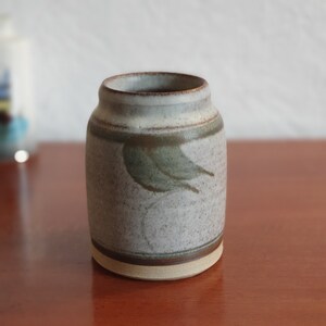 May include: A small, white ceramic vase with a brown and green leaf design. The vase has a speckled glaze and a rounded shape.