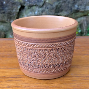 May include: A brown ceramic planter with a textured design. The planter has a wide, round opening and a slightly flared lip.