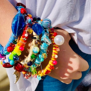 May include: A colorful stack of bracelets with beads, fabric, and wire. The bracelets are in shades of blue, red, yellow, green, and white.
