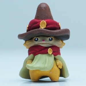 May include: A whimsical figurine of a creature wearing a brown wizard hat with gold accents and a red scarf. The figure has a yellow body, green cape, and is set against a light blue background.