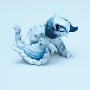 May include: A small, white and blue fantasy creature figurine. The figurine has a dragon-like appearance with horns, spikes, and a flowing tail. The creature is painted with shades of blue and white, giving it a whimsical look. The figurine is set against a plain white background.