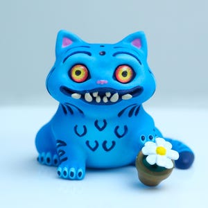 Puede incluir: Una caprichosa figura de gato azul con grandes ojos amarillos, una nariz rosa y una sonrisa dentuda. El gato tiene marcas negras y sostiene una pequeña maceta con una flor blanca y amarilla. Un artículo decorativo único.