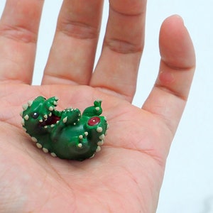 May include: A small green dinosaur figurine with white spots and red eyes. The dinosaur is lying on its back with its mouth open.