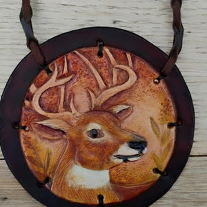May include: A circular leather pendant featuring a hand-tooled and painted deer head with antlers in shades of brown, tan, and white. The piece is framed by a dark brown leather border with black stitching and hangs from a braided leather cord.