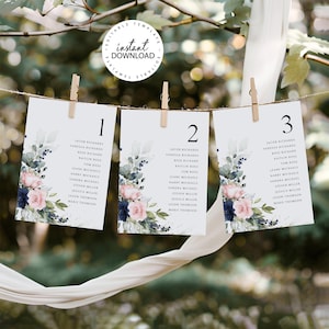 May include: Three white cards with floral designs and numbers 1, 2, and 3 printed on them. The cards are hanging from a string with clothespins. Each card lists names for a wedding seating chart.