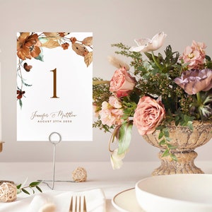 May include: A white wedding table number card with a brown floral design and the number "1" in gold. The card also includes the names "Jennifer & Matthew" and the date "August 27th 2020".