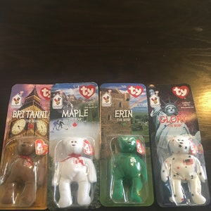 May include: Four Ty Beanie Babies, each representing a different country. The bears are named Britanni, Maple, Erin, and Glory. Each bear is wearing a small tag with the Ty logo and the bear's name. The bears are displayed on a wooden surface.