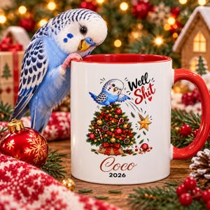 May include: A white ceramic mug with a red interior and handle, featuring a graphic of a blue budgie perched on a decorated Christmas tree with the text Well Shit, the name Coco, and the year 2026 printed on the front surface of the mug.