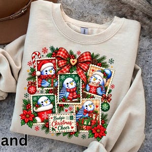 May include: A sand-coloured crewneck sweatshirt featuring a festive graphic print of four blue budgies in postage stamp frames, decorated with a red plaid bow, holly, poinsettias, and ornaments. The text reads Budgie Christmas Cheer.